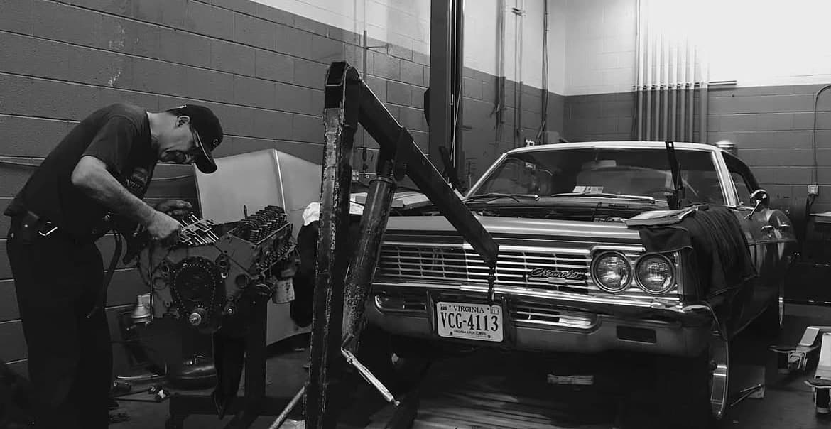 A mechanic working on an engine block next to a 1966 Chevrolet Impala in our Purcellville shop